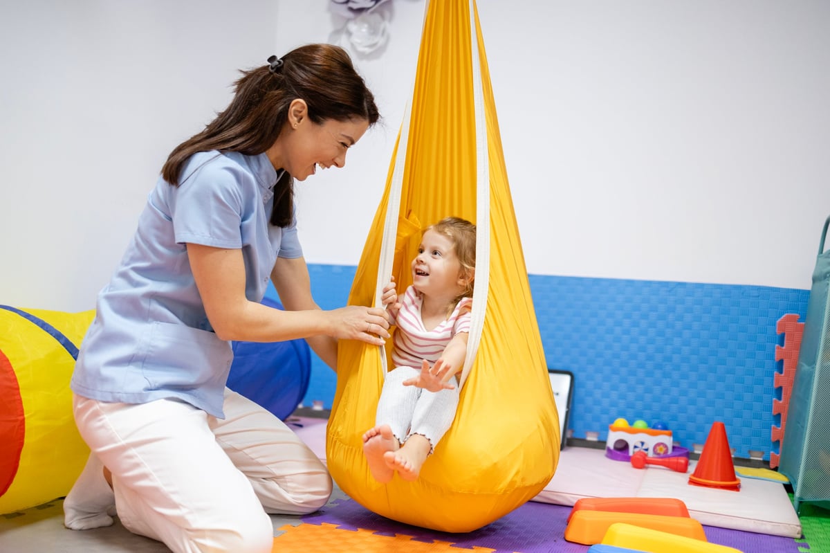 Therapist supporting a child on a sensory swing in a bright therapy gym