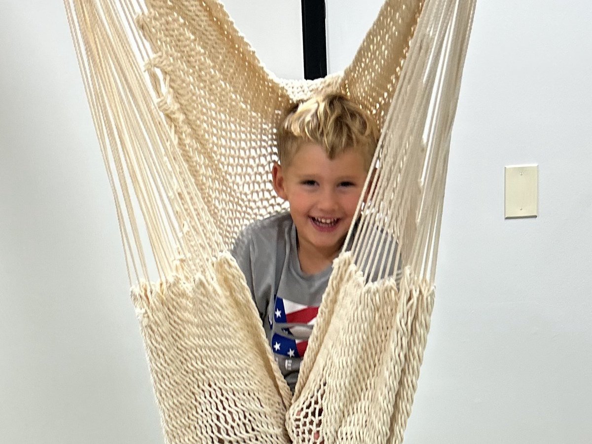 Blond child smiling while sitting in a beige mesh hammock swing, calming sensory input in the sensory gym at New Heights Therapy