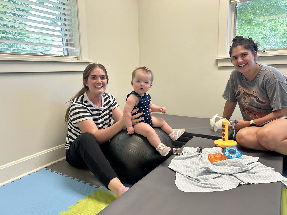 Infant sitting balance practice on a therapy ball with two pediatric physical therapists in a bright therapy room