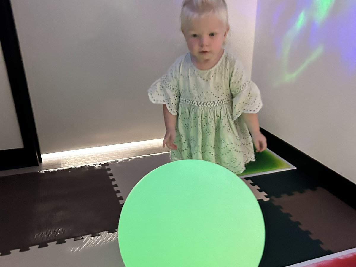 Toddler in a green dress watching a large glowing green light-up ball in the sensory room at New Heights Therapy