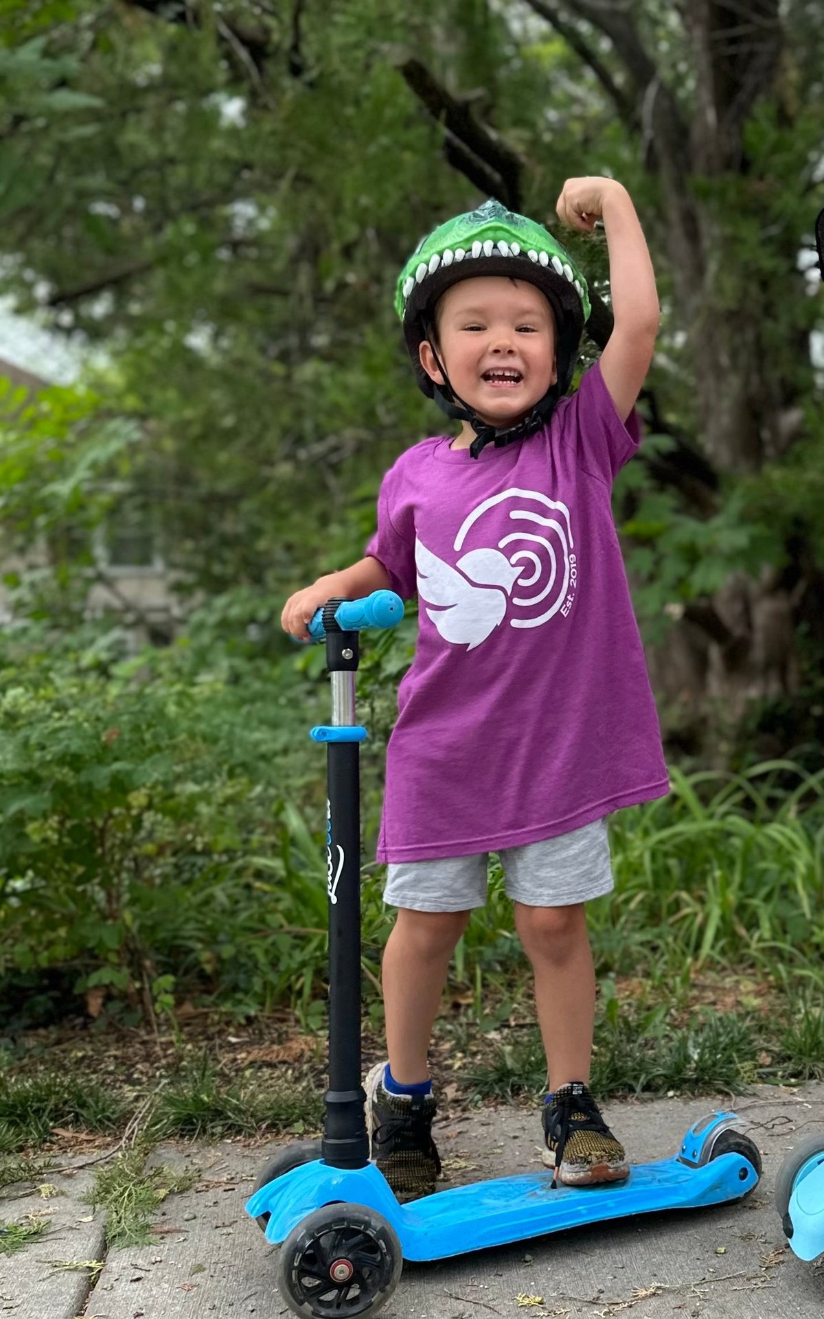 Young child in a purple shirt and green helmet standing on a blue three-wheeled scooter with a fist raised outdoors — the kind of confidence pediatric physical therapy builds at New Heights Therapy in Lawrence and Topeka, Kansas