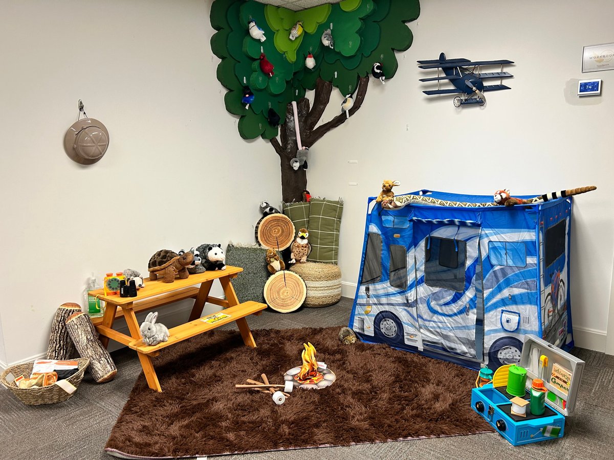 Nature nook at New Heights Therapy in Lawrence, Kansas, with a green tree wall mural, plush forest animals like raccoons, owls and a fox, a wooden picnic table, log cushions, a toy campfire on a brown rug and a blue campervan play tent for pediatric speech and occupational therapy