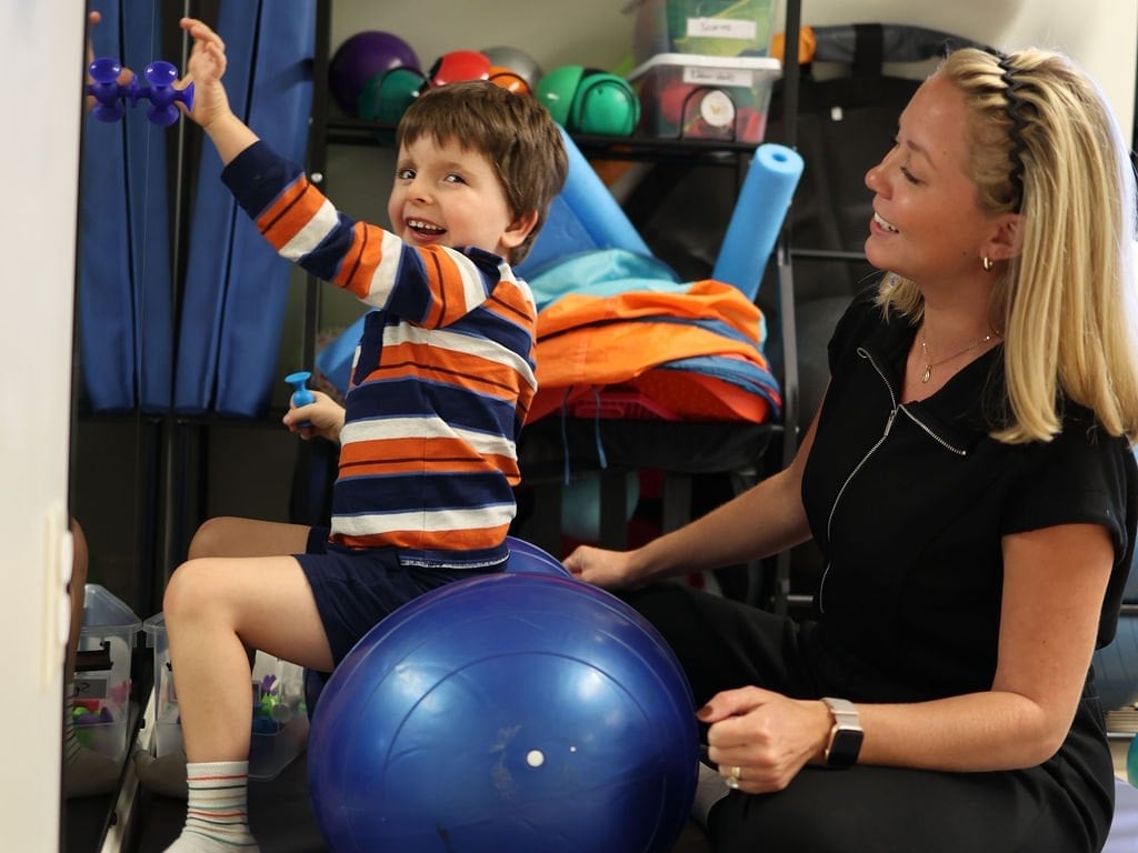 Child on a therapy ball working on core strength and reaching during physical therapy, while the therapist supervises