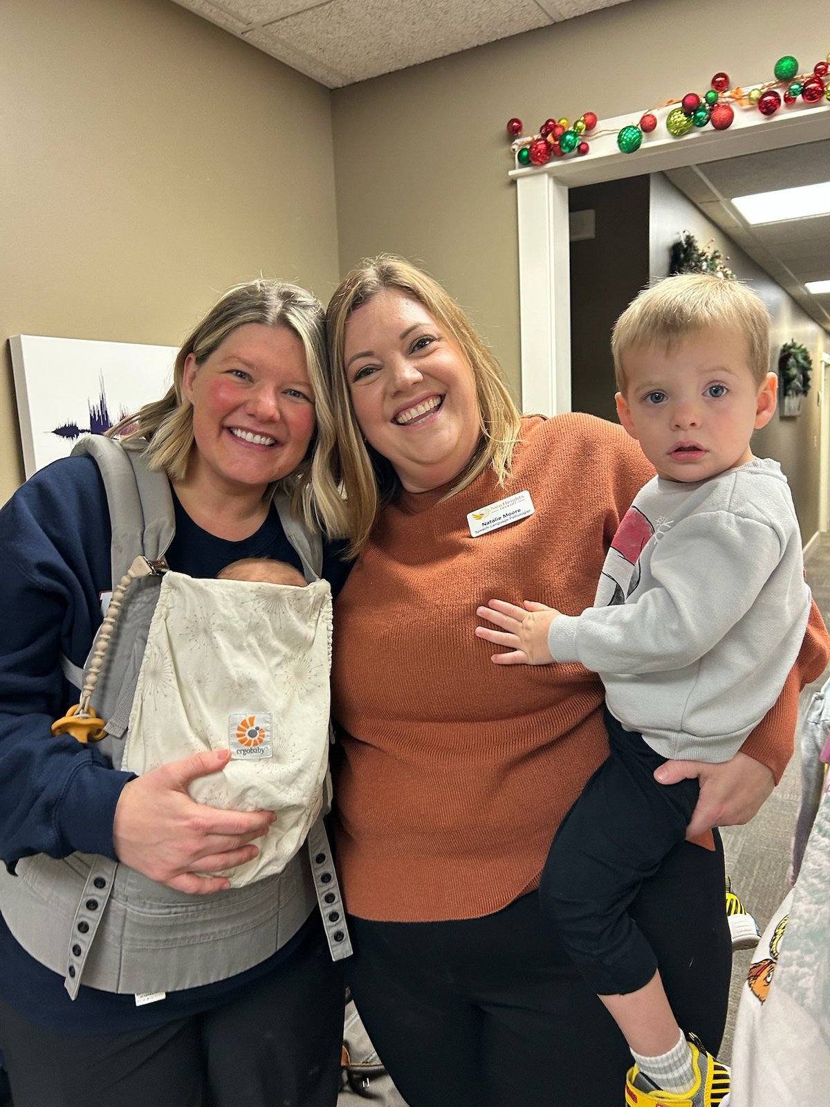 A mother wearing her baby in a carrier stands smiling beside a New Heights Therapy therapist holding a toddler — the parent-and-therapist partnership at the heart of every care plan