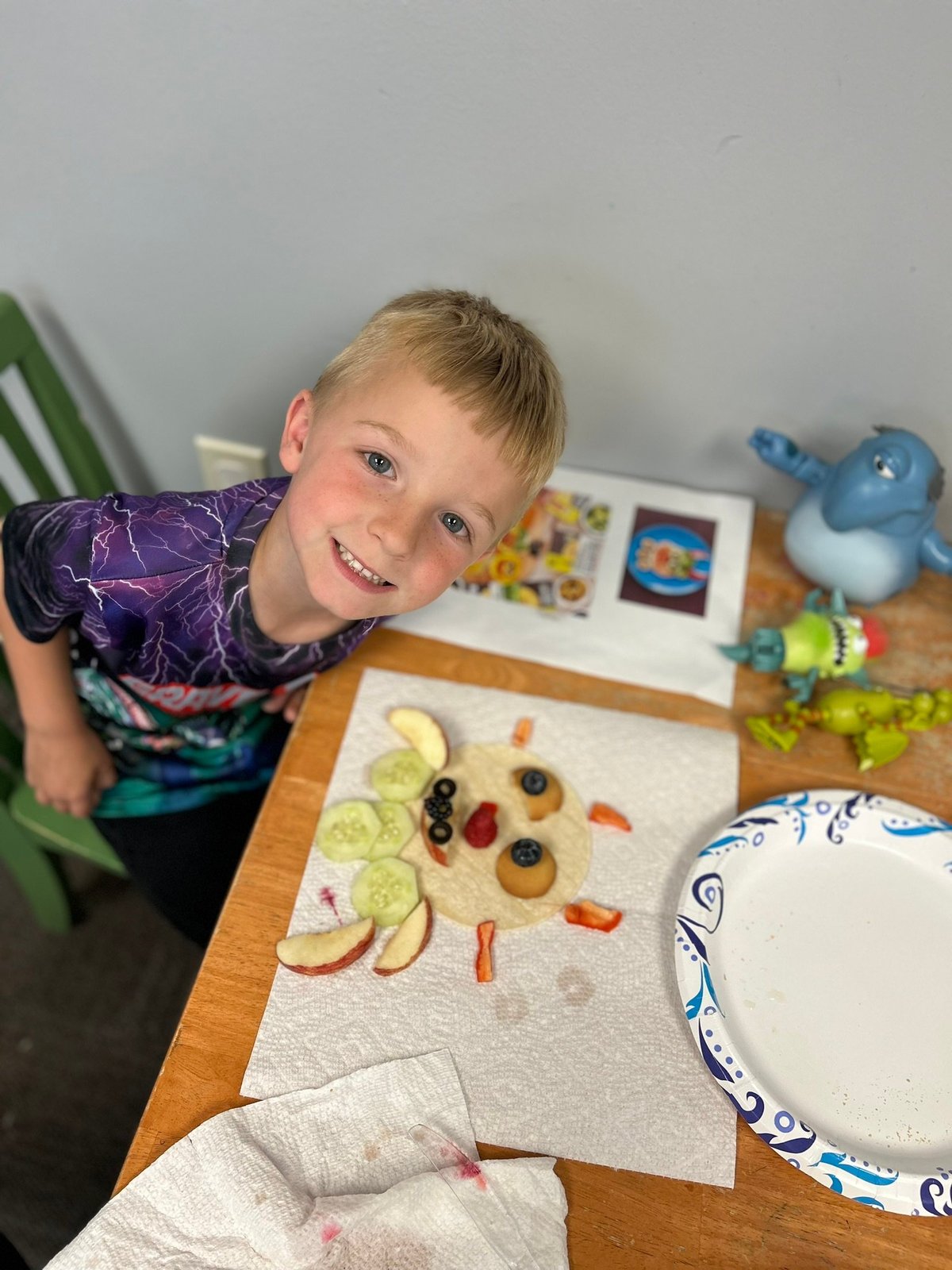 Young boy smiling at a table where food has been arranged into a funny face — playful pediatric feeding therapy at New Heights Therapy in Lawrence and Topeka, Kansas