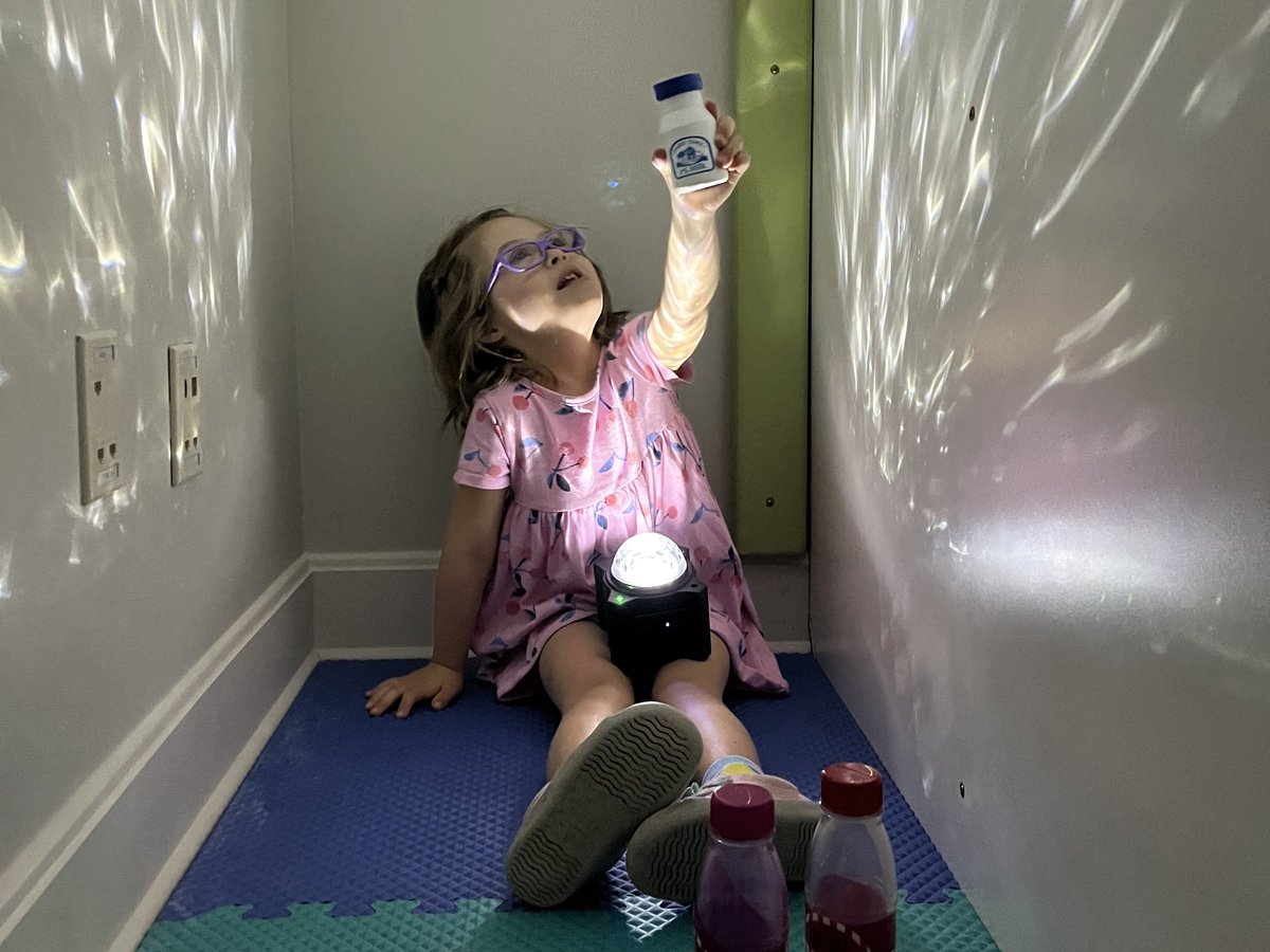 Child in a pink dress and glasses holding an illuminated bottle under the sensory room light projector, with light patterns on the walls