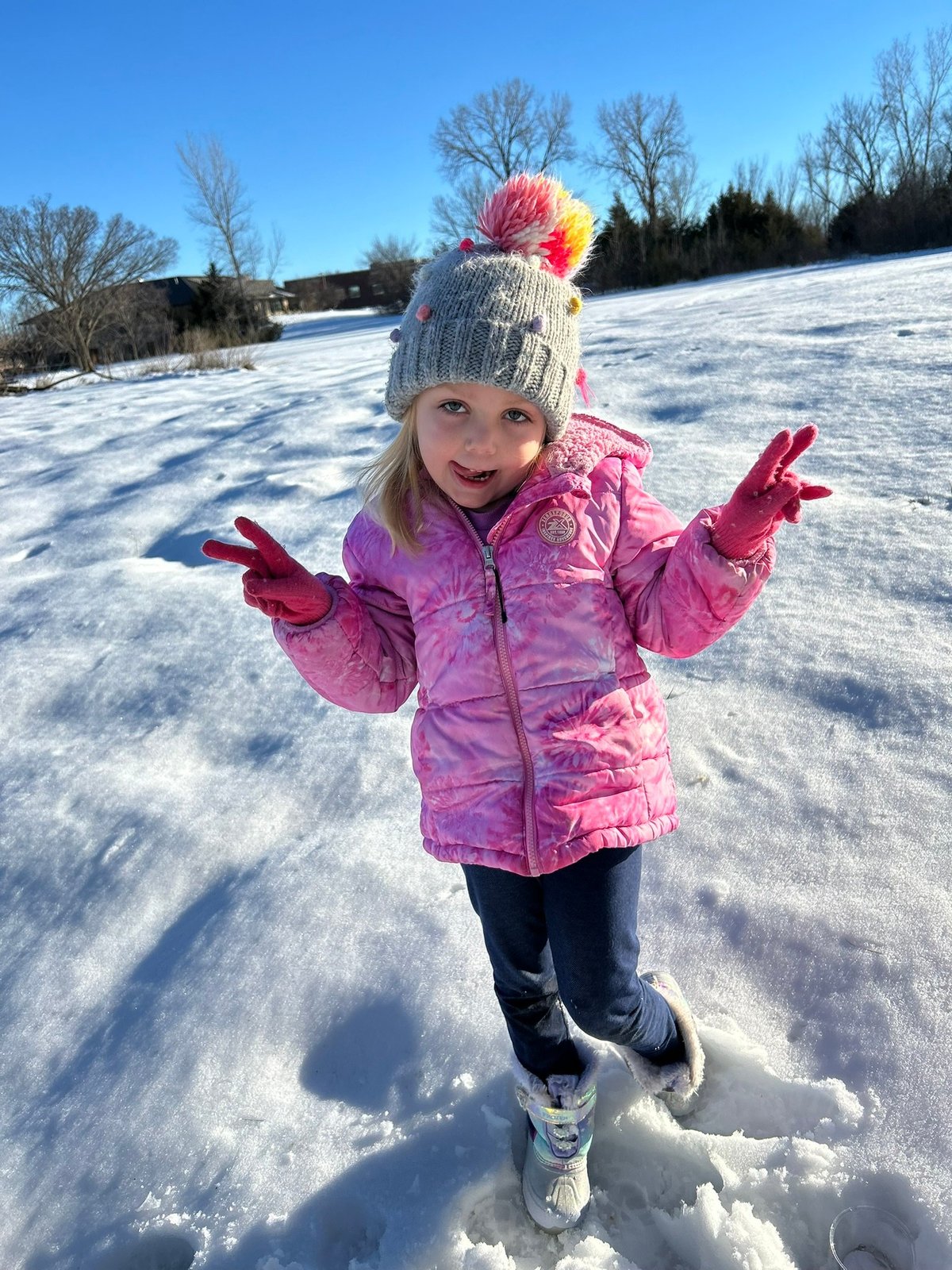 Young girl smiling with her arms raised in a snowy field, wearing a pink jacket and colorful pom-pom hat