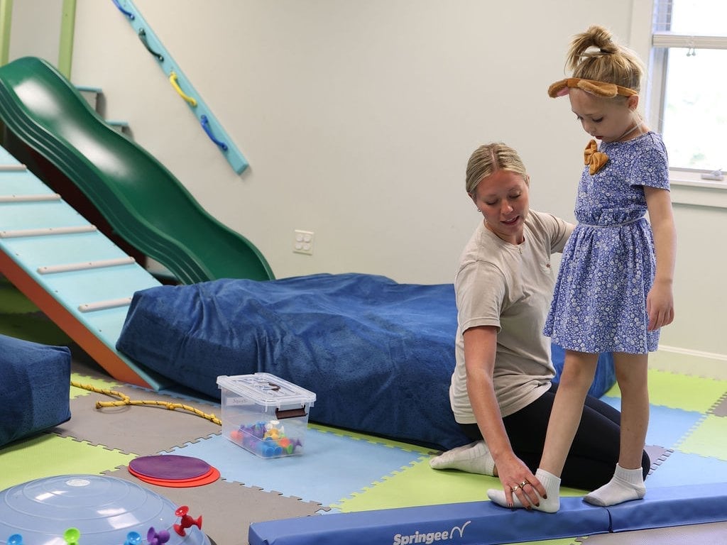 Child walking a balance beam during pediatric physical therapy, with a slide, blue tunnel and colorful mats in the clinic gym