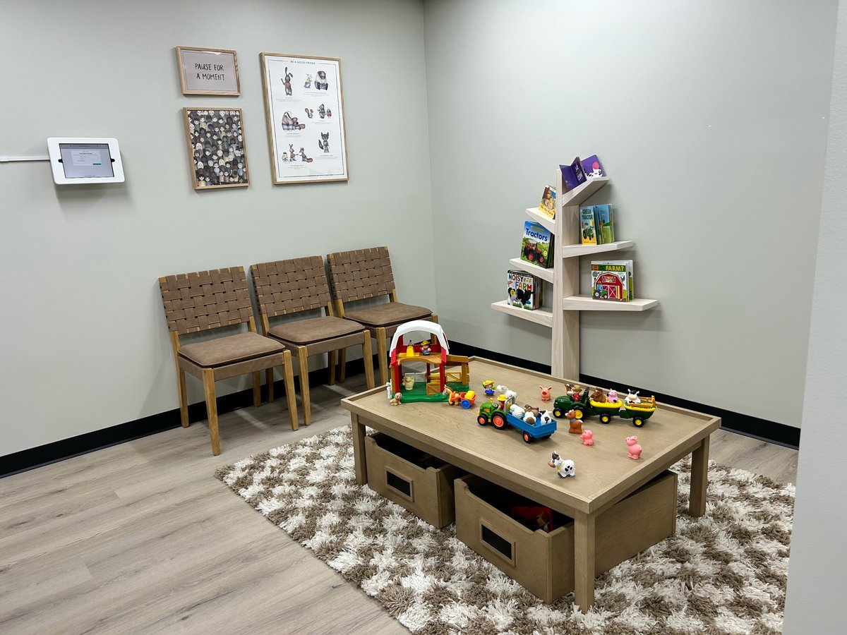 A childs playroom with wooden chairs, a play table with toys, geometric rug, wall art, and shelving unit with books and decorations