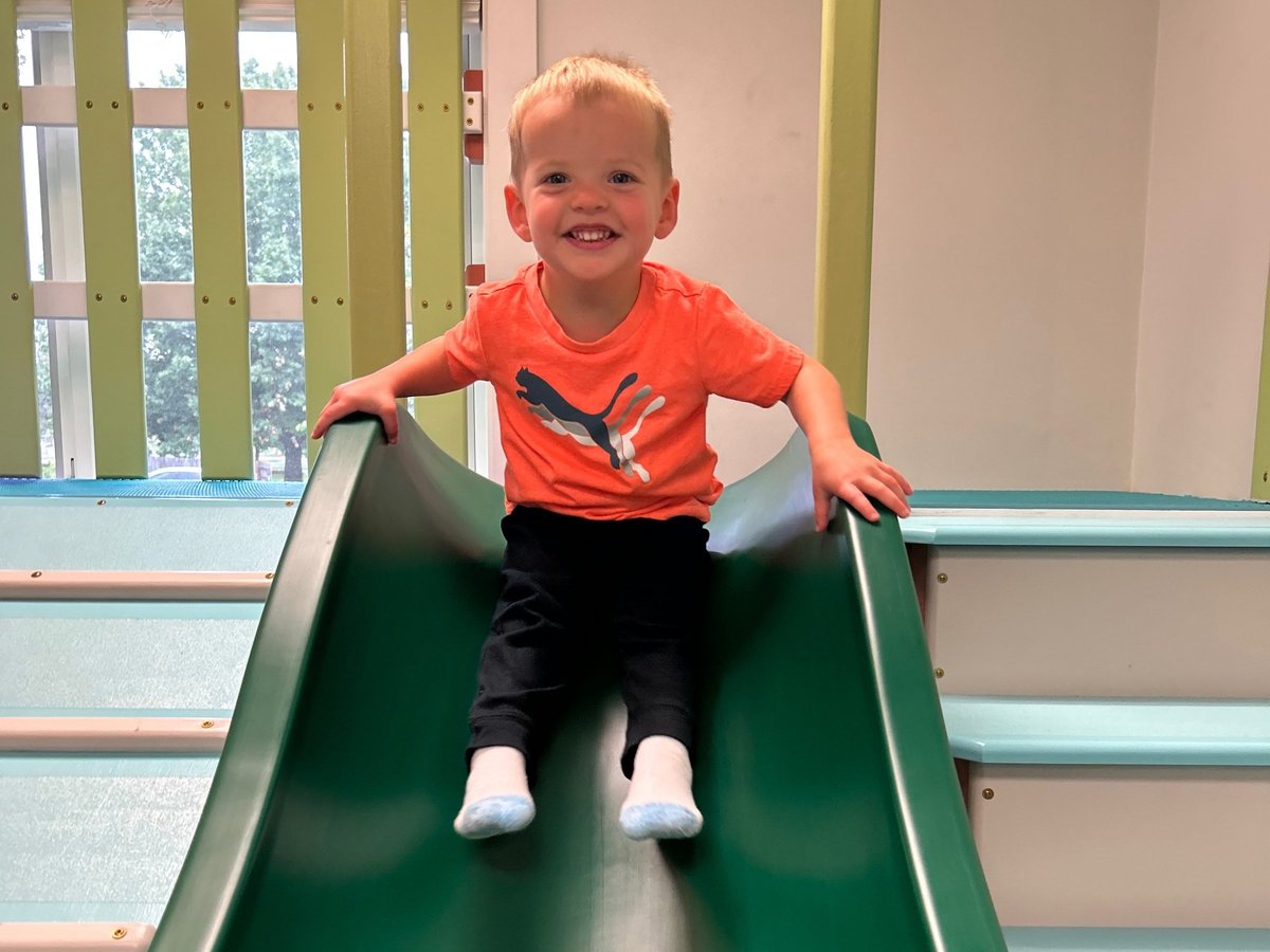 Smiling toddler in an orange shirt sitting at the top of the green slide in the sensory gym, pediatric occupational therapy at New Heights Therapy