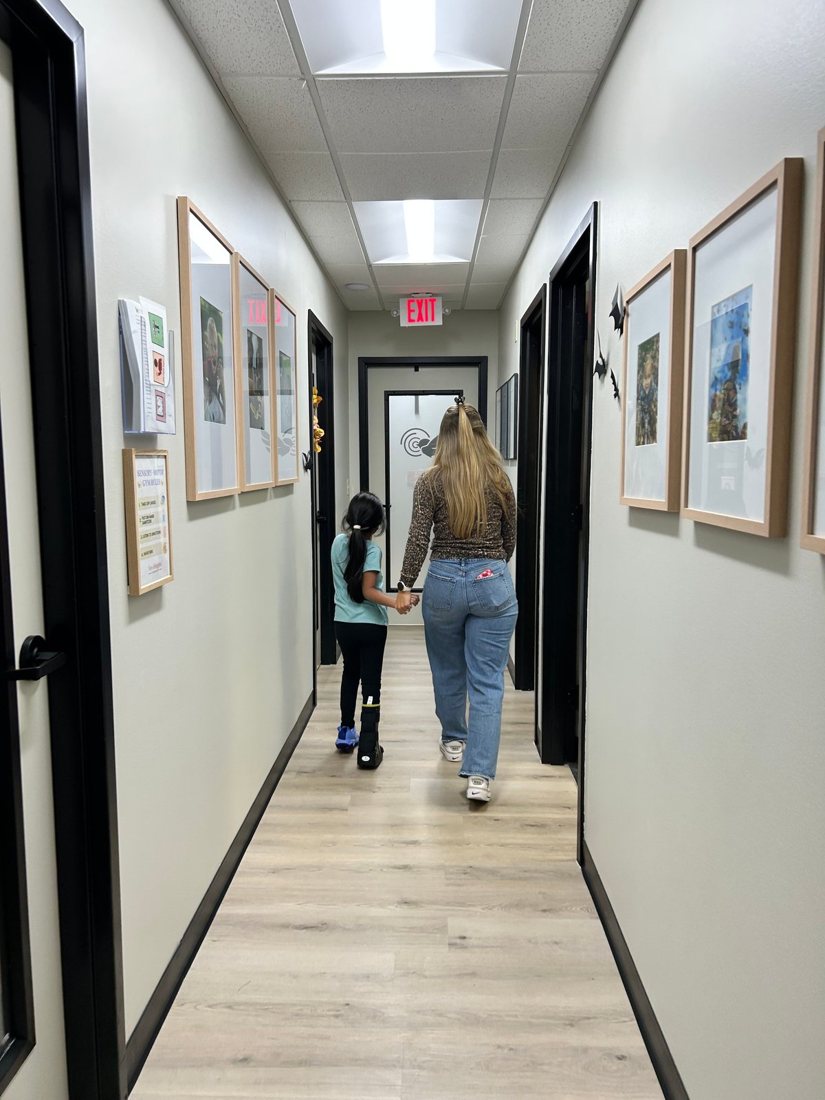 Two people walking down a narrow hallway lined with framed artwork on white walls and black doors