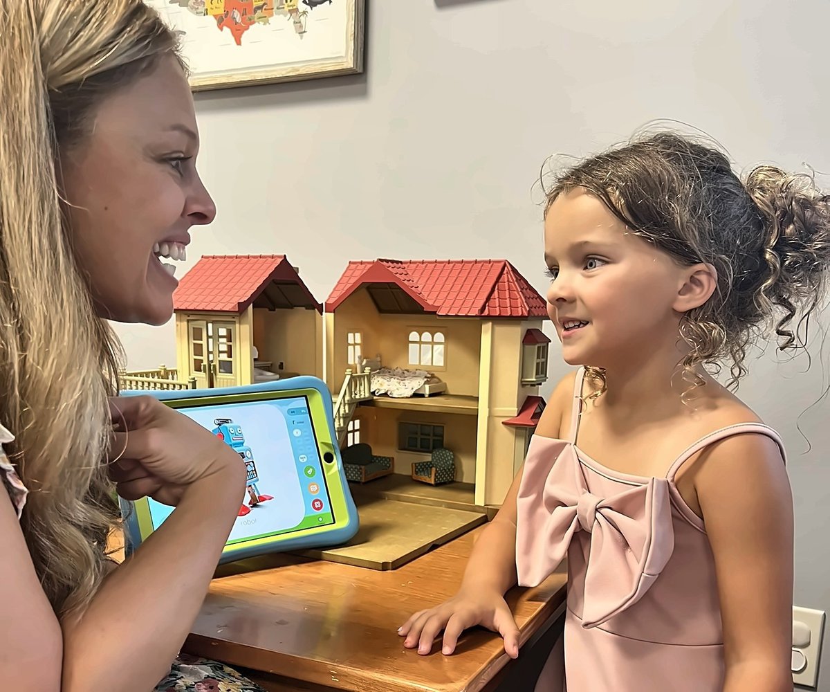Speech therapist and a young girl using a tablet with a dollhouse, building language and following directions through play at New Heights Therapy