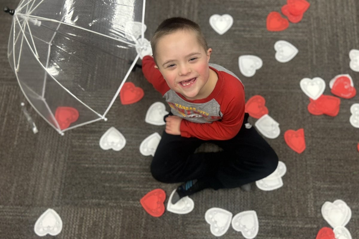 Young child in red shirt sitting on gray floor surrounded by red and white paper hearts, holding a decorative umbrella.