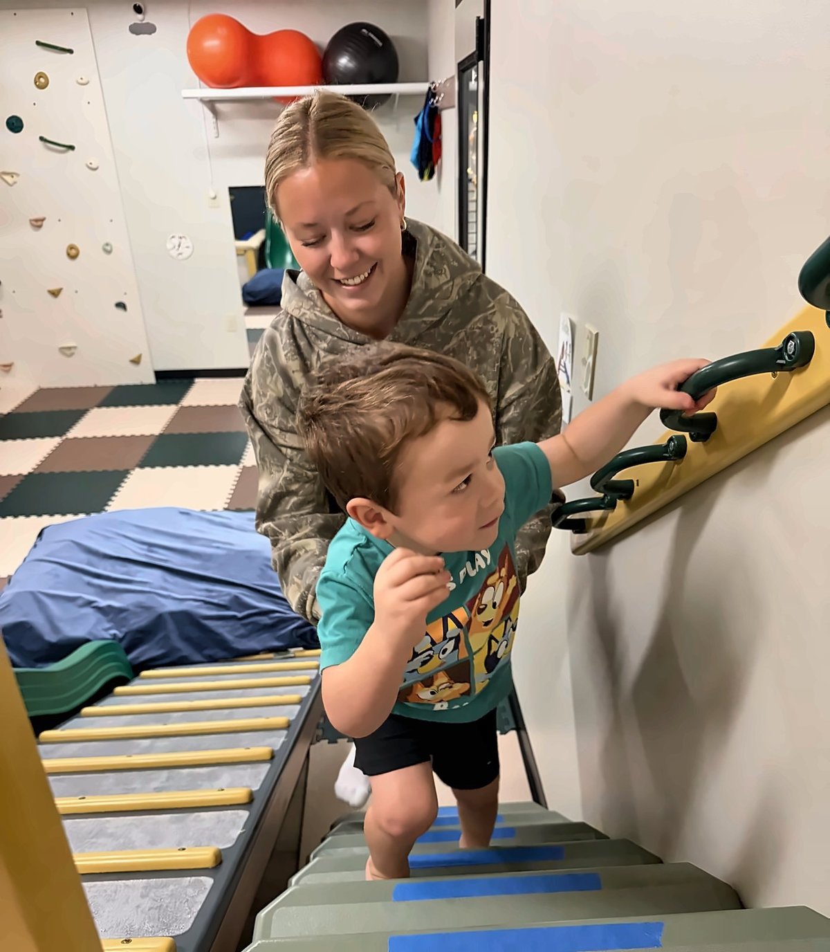 Pediatric physical therapist guiding a toddler across a yellow balance beam in the sensory gym — building balance and coordination at New Heights Therapy in Lawrence and Topeka, Kansas