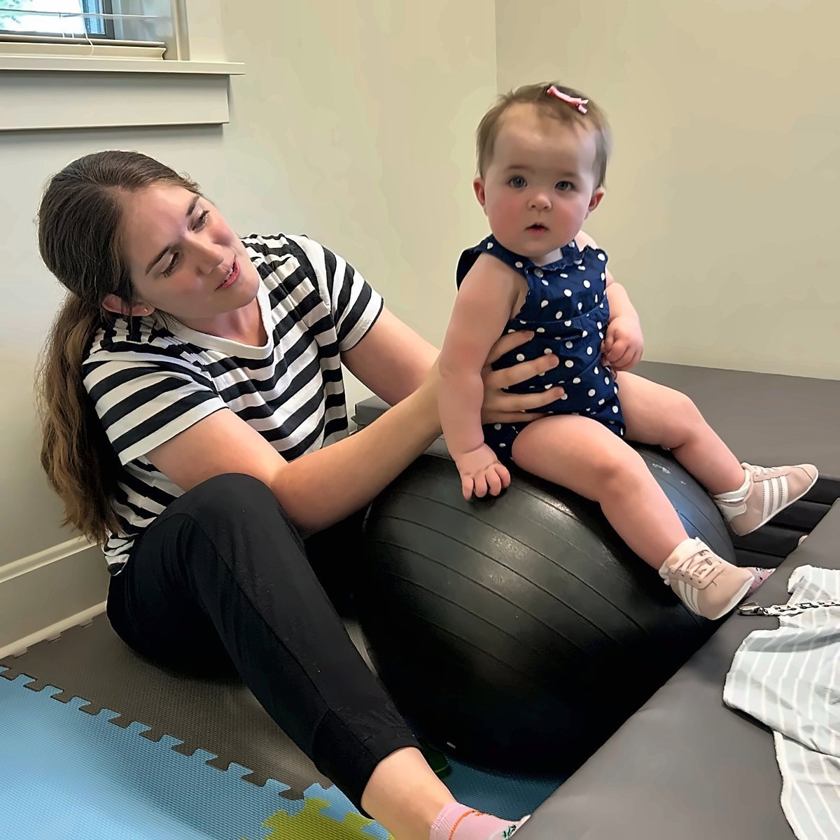 Woman in striped shirt sitting on exercise ball with toddler in navy polka dot outfit on a padded floor