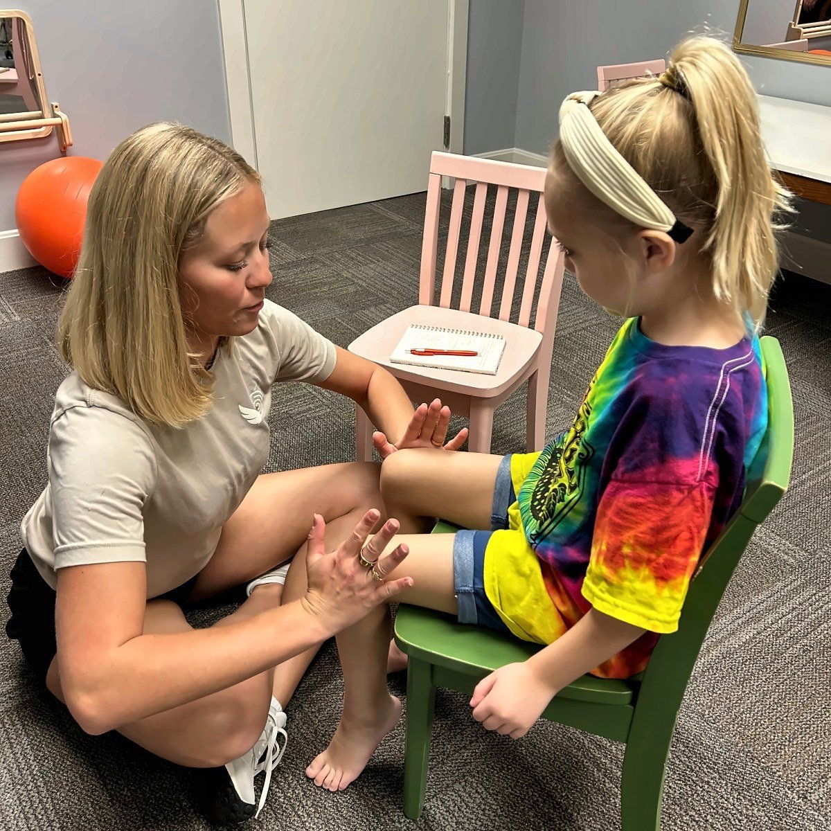 Pediatric physical therapist sitting on the floor working with a young girl seated on a chair, guiding her hand during a balance and strengthening session at New Heights Therapy in Kansas