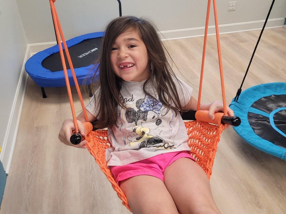 Young girl smiling on a net swing with orange ropes, vestibular sensory input in the New Heights Therapy sensory gym