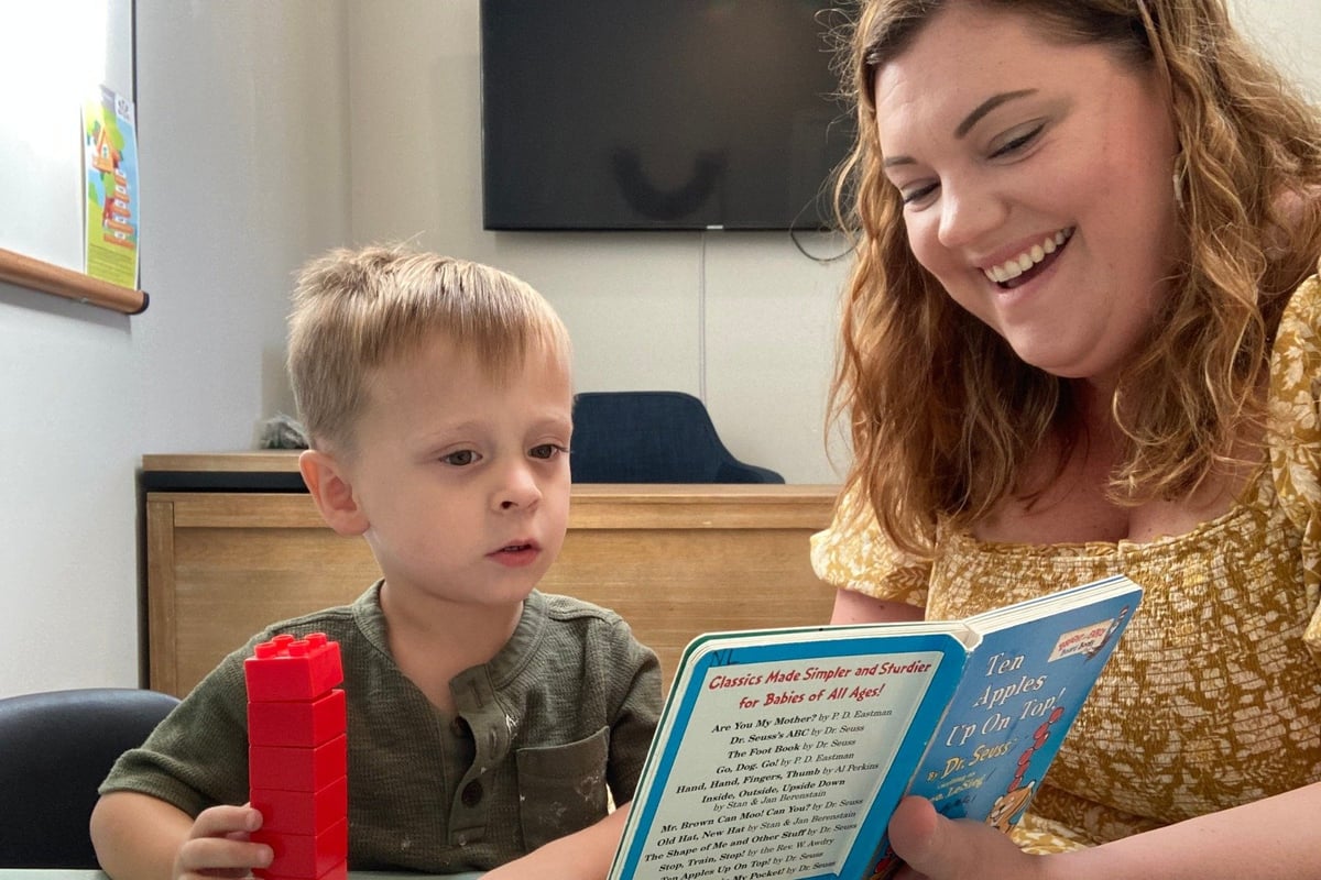 Woman reading a children's book to a young toddler boy in an indoor setting.
