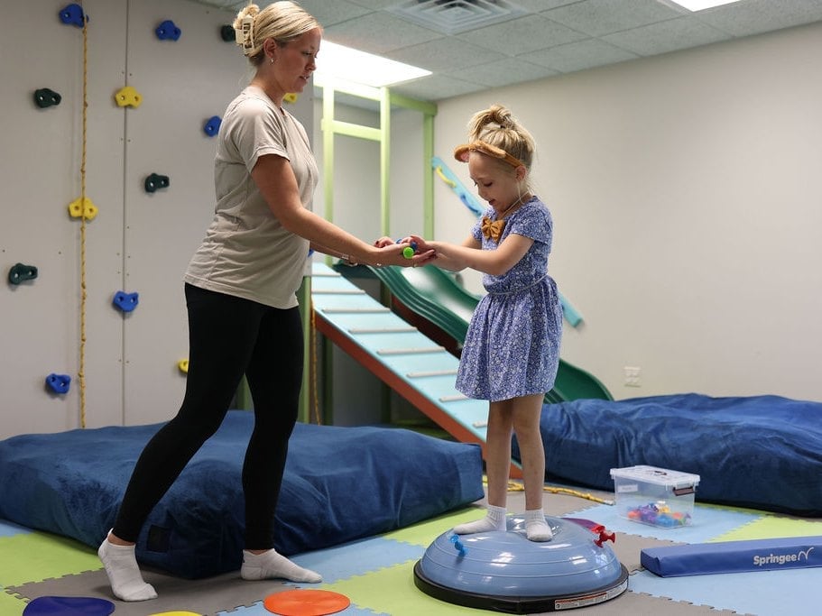 Child standing on a bosu ball for balance practice in pediatric physical therapy, a therapist supporting each step in the colorful therapy gym
