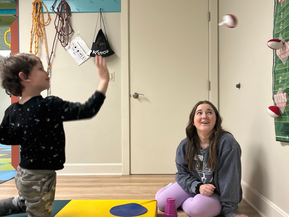 Young child tossing a ball to a therapist sitting on the floor, practising hand-eye coordination during occupational therapy at New Heights Therapy