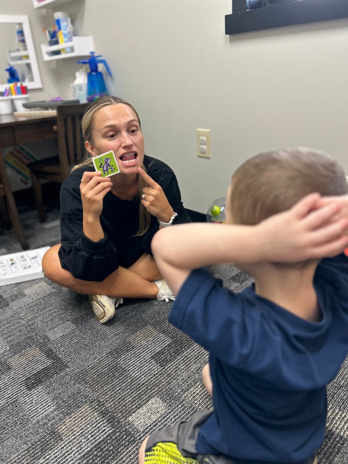 Speech therapist holding up a picture card and modeling a mouth position while a young child watches during articulation therapy