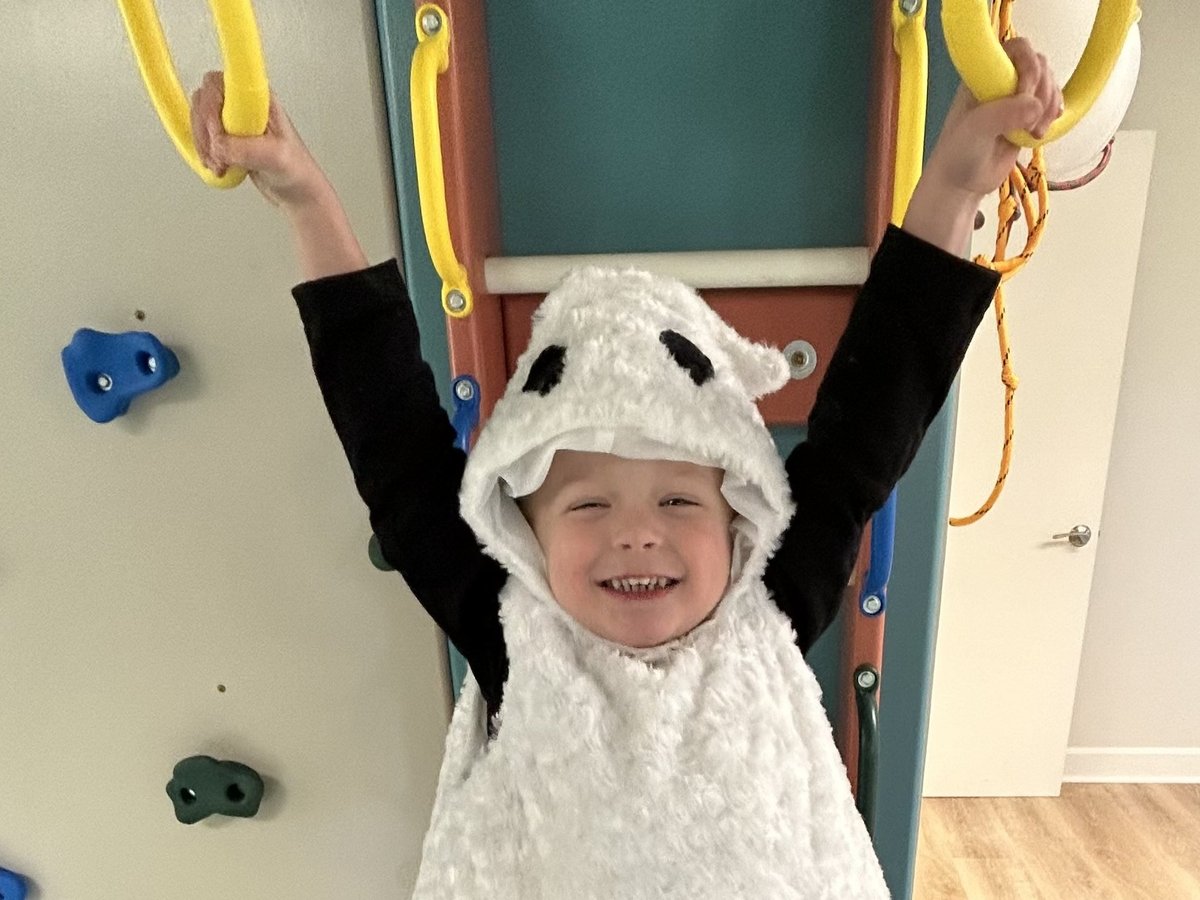 Smiling child in a sheep costume holding yellow gymnastics rings with raised arms, building upper body strength in occupational therapy