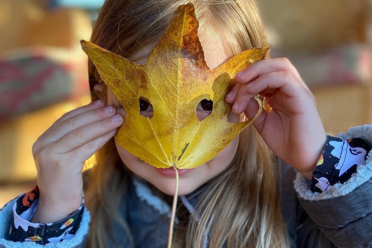Child holding a large yellow leaf up to their face like a mask, with blonde hair visible and blurred outdoor background.