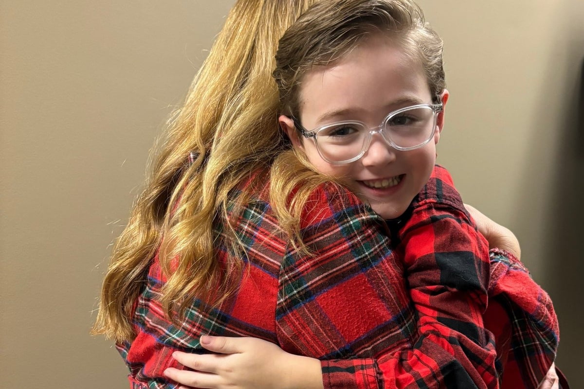 Child wearing clear-framed glasses and red plaid shirt, smiling at camera against neutral background.