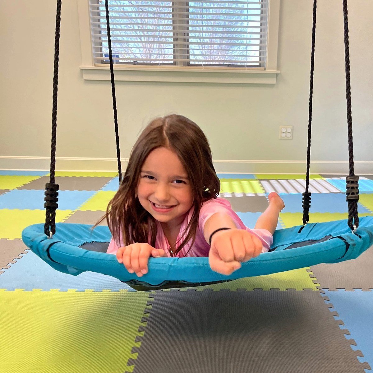 Young girl smiling as she lies on a blue platform swing in the sensory gym at New Heights Therapy, a pediatric occupational therapy clinic in Lawrence, Kansas