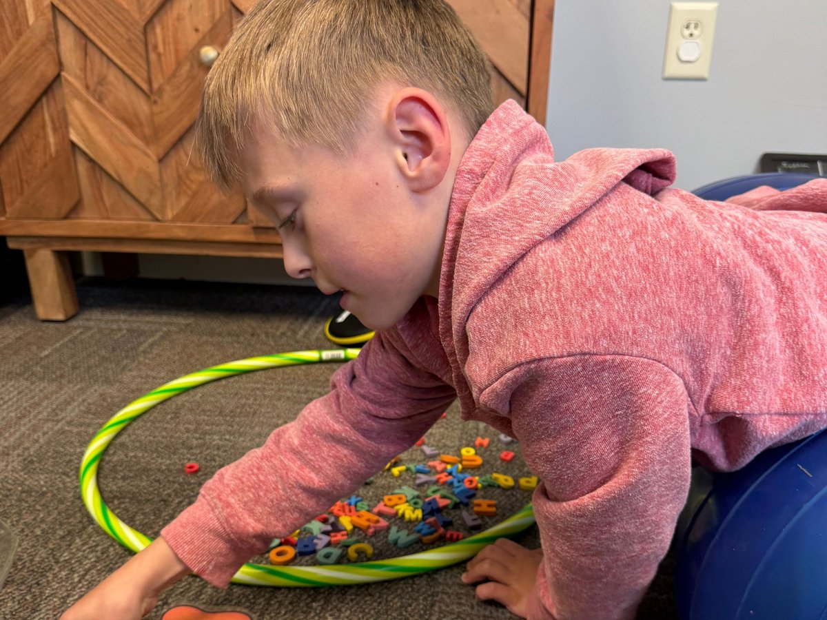 Young child in a pink hoodie matching letters while lying over a therapy ball, building core strength during occupational therapy