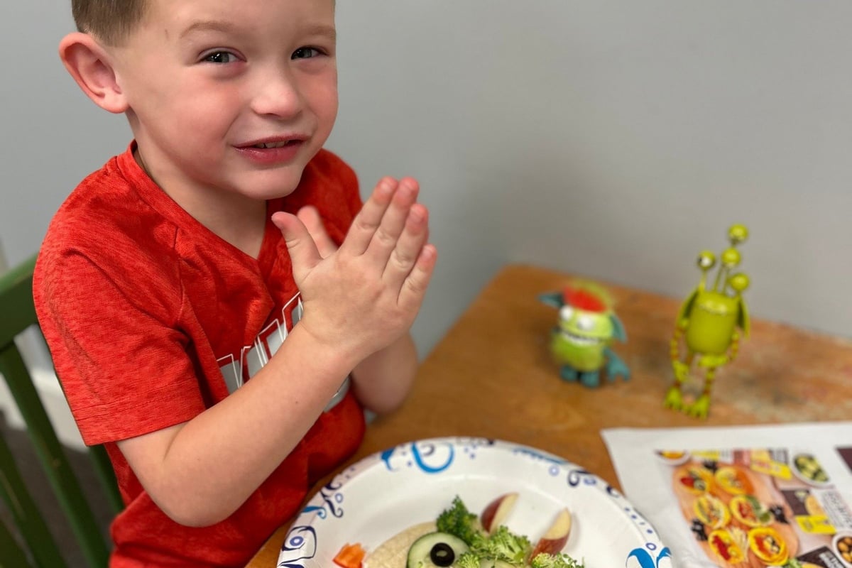 Young child in red shirt clapping hands at table with decorated plate and toy figures.