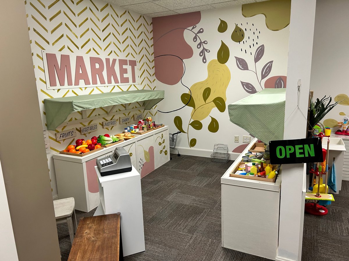 Colorful pretend play market area with a toy kitchen, displays of play food and decorative fruit murals on the wall