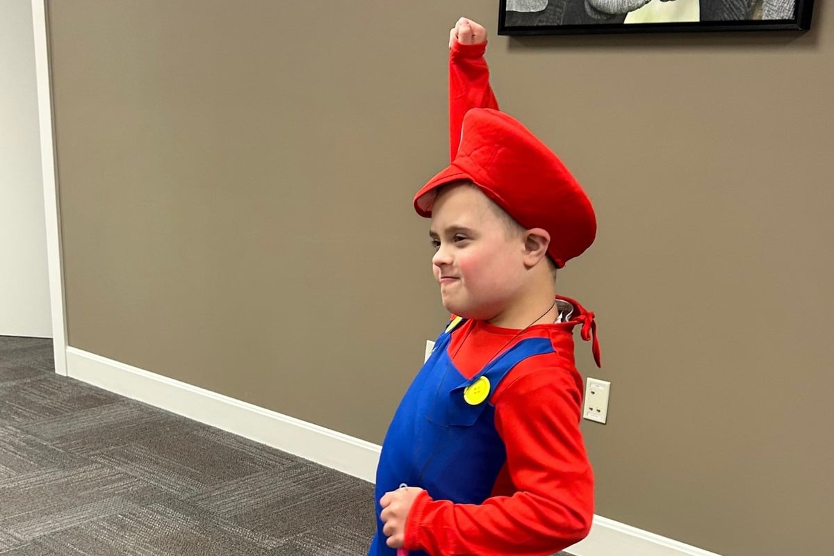 Child wearing Mario costume with red hat, blue overalls, and red shirt posing in an office setting.