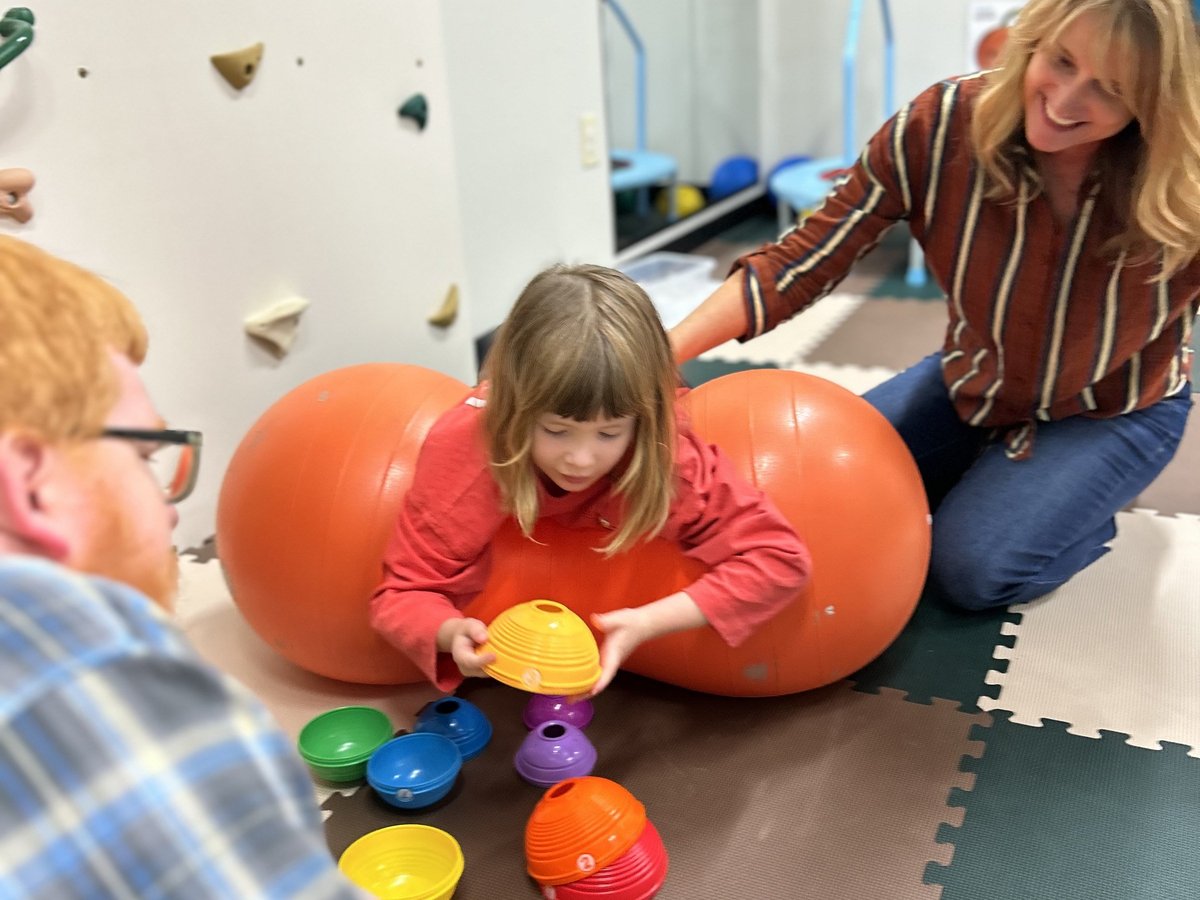 Child in a red jacket stacking colourful nesting cups on a peanut ball while a therapist supervises in the clinic classroom