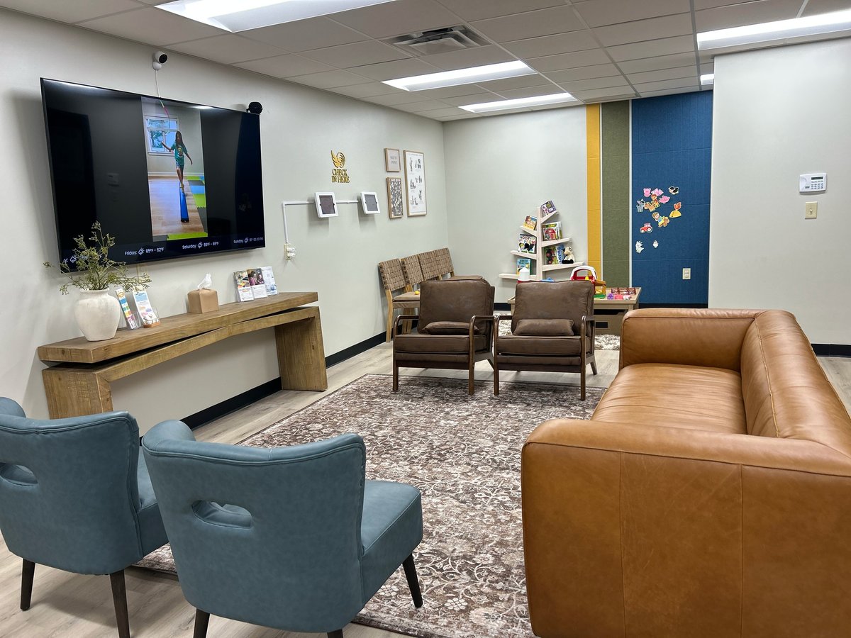 Modern office lounge with brown leather sofa, blue armchairs, wall-mounted TV, wooden shelving, and decorative blue accent wall