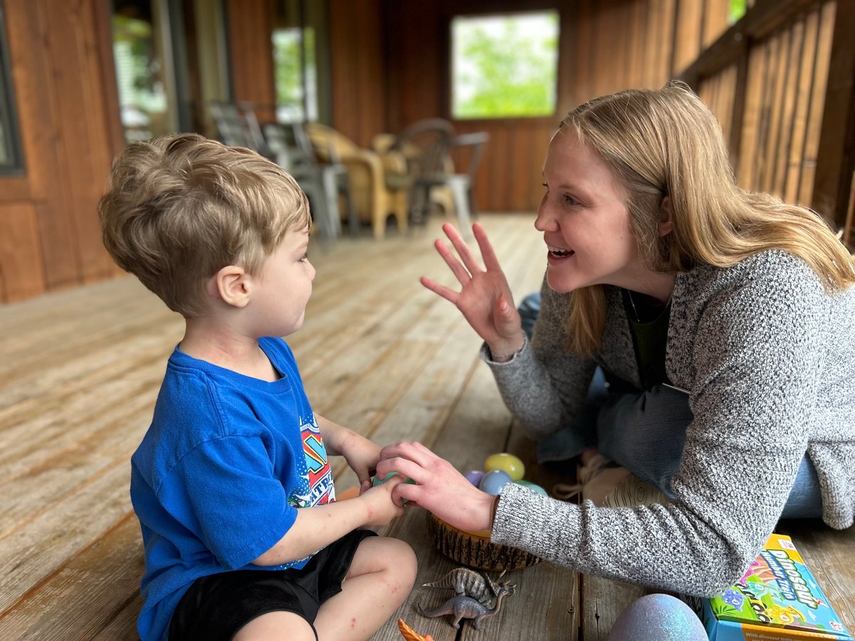 Pediatric therapist kneeling face-to-face with a young boy in a blue shirt on a wooden porch, playing together during a session with New Heights Therapy
