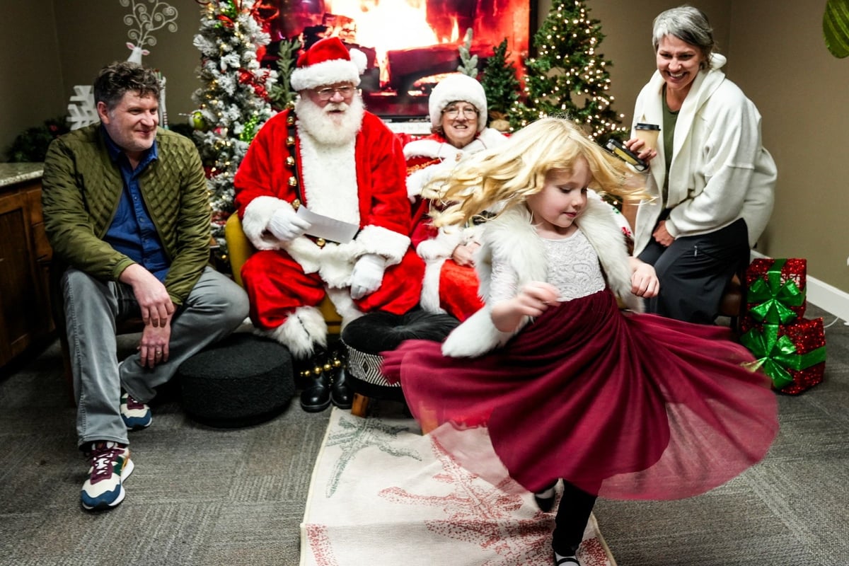 Young girl twirling in burgundy skirt with Santa and family members during holiday celebration indoors.