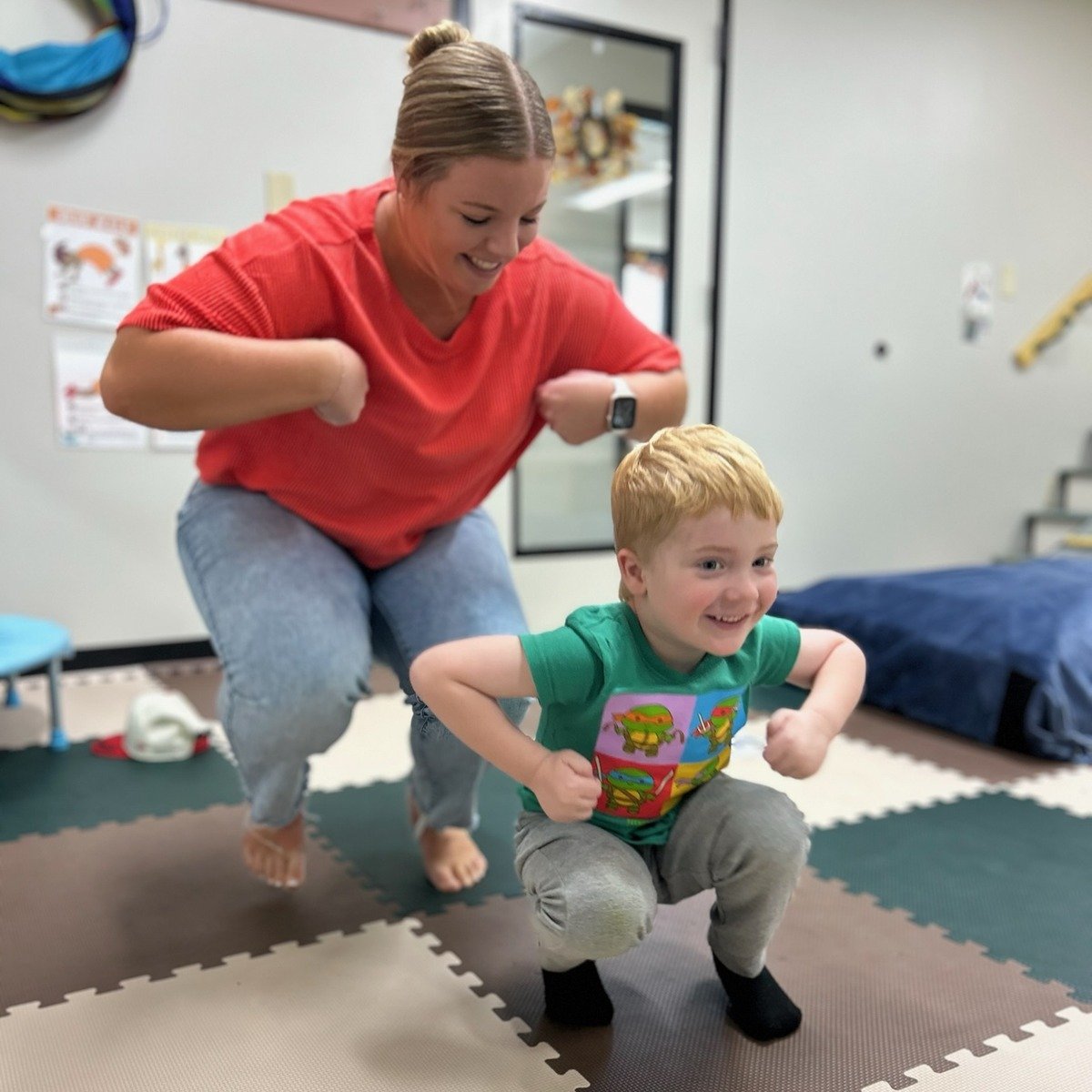 Woman in red shirt helping young child in green shirt jump on foam play mats in a therapy or daycare setting