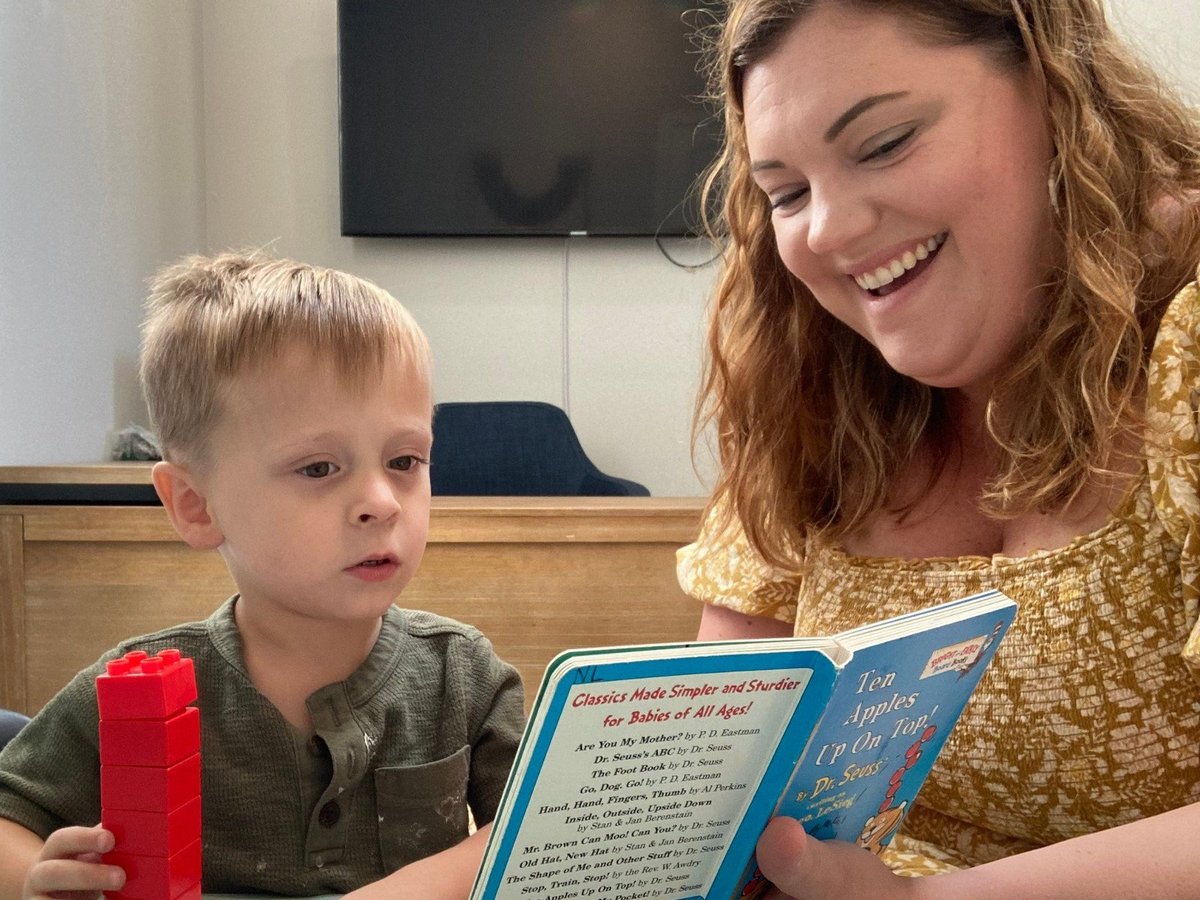 Woman reading a childrens book to a young boy while holding a red toy in an indoor living room setting