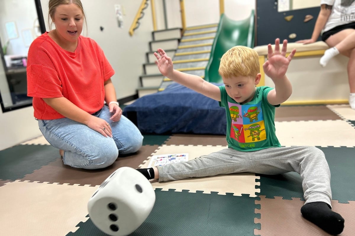 A caregiver in a red shirt watches a young child in a green shirt playing with a large white die on a foam mat in an indoor gym or play space.