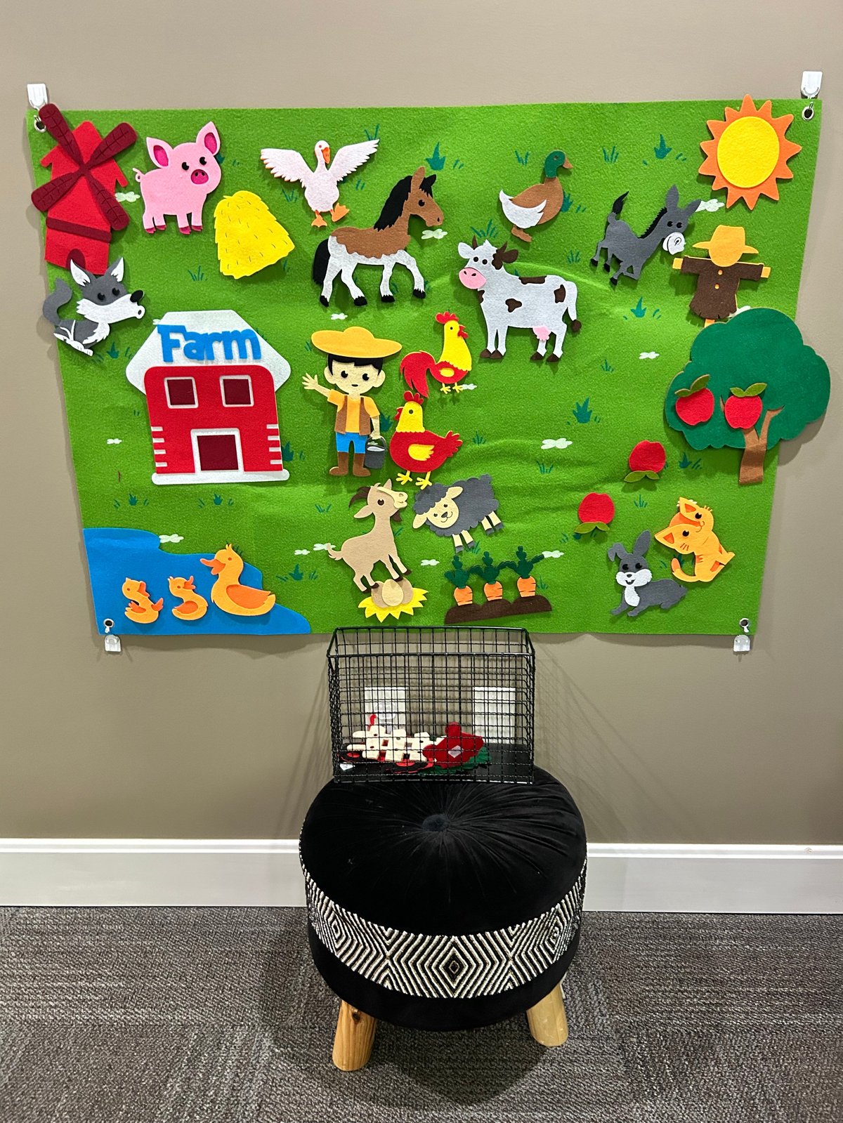 Felt board mounted on wall with colorful cutout animals, barn, farmer figure, and sun above a black storage basket with wooden legs