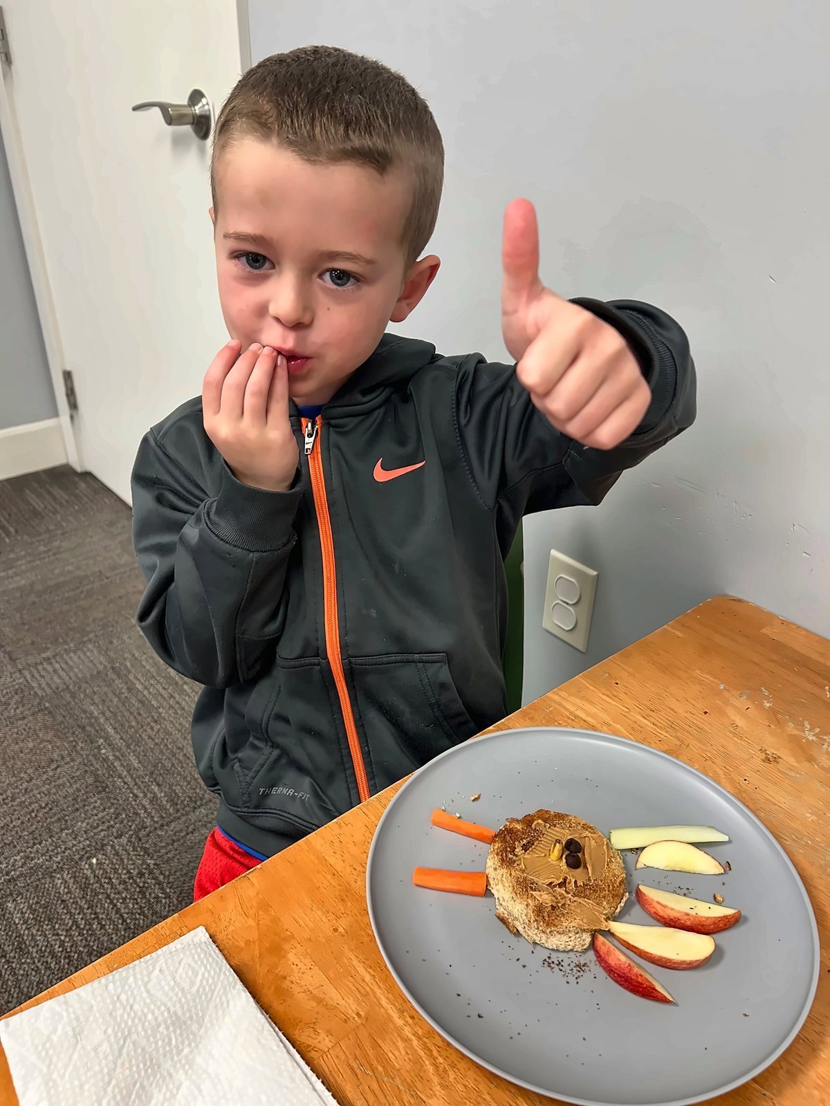 Young child in a black jacket giving a thumbs up while eating, with a plate of carrot sticks and apple slices — a win during pediatric feeding therapy at New Heights Therapy in Kansas
