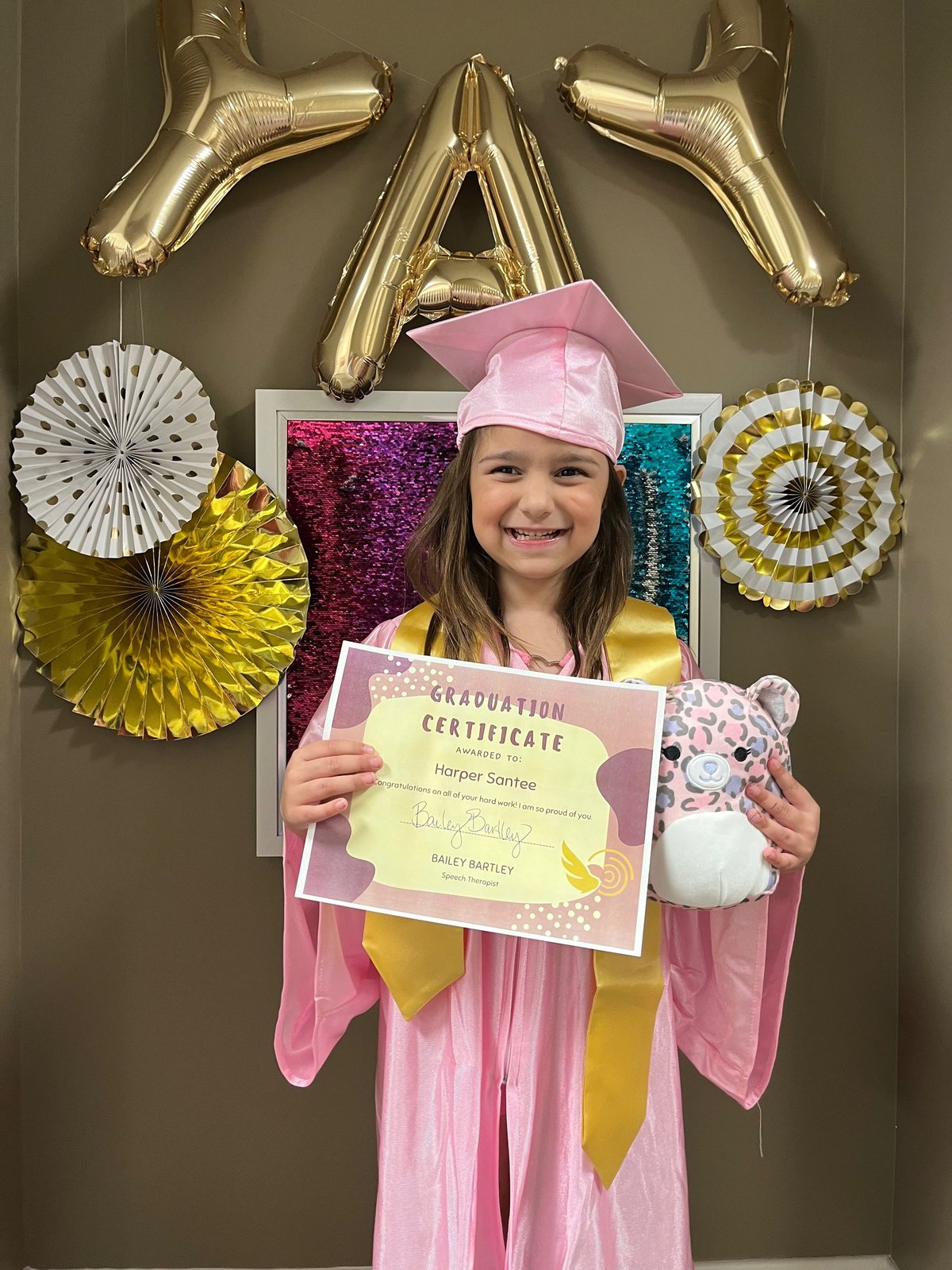 Speech therapy graduation day in Lawrence, Kansas — a young girl in a pink gown holding her certificate in front of balloons at New Heights Therapy