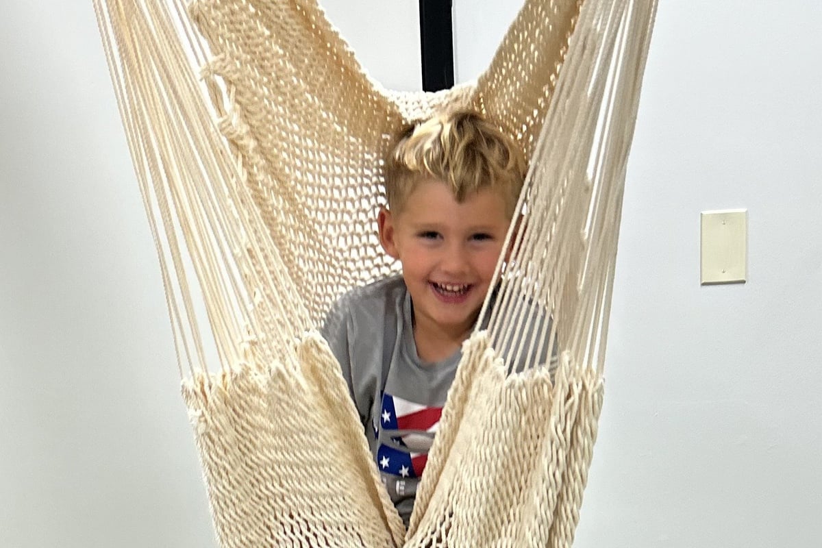 Young child smiling in a beige rope hammock, wearing a blue patterned shirt against a white wall.