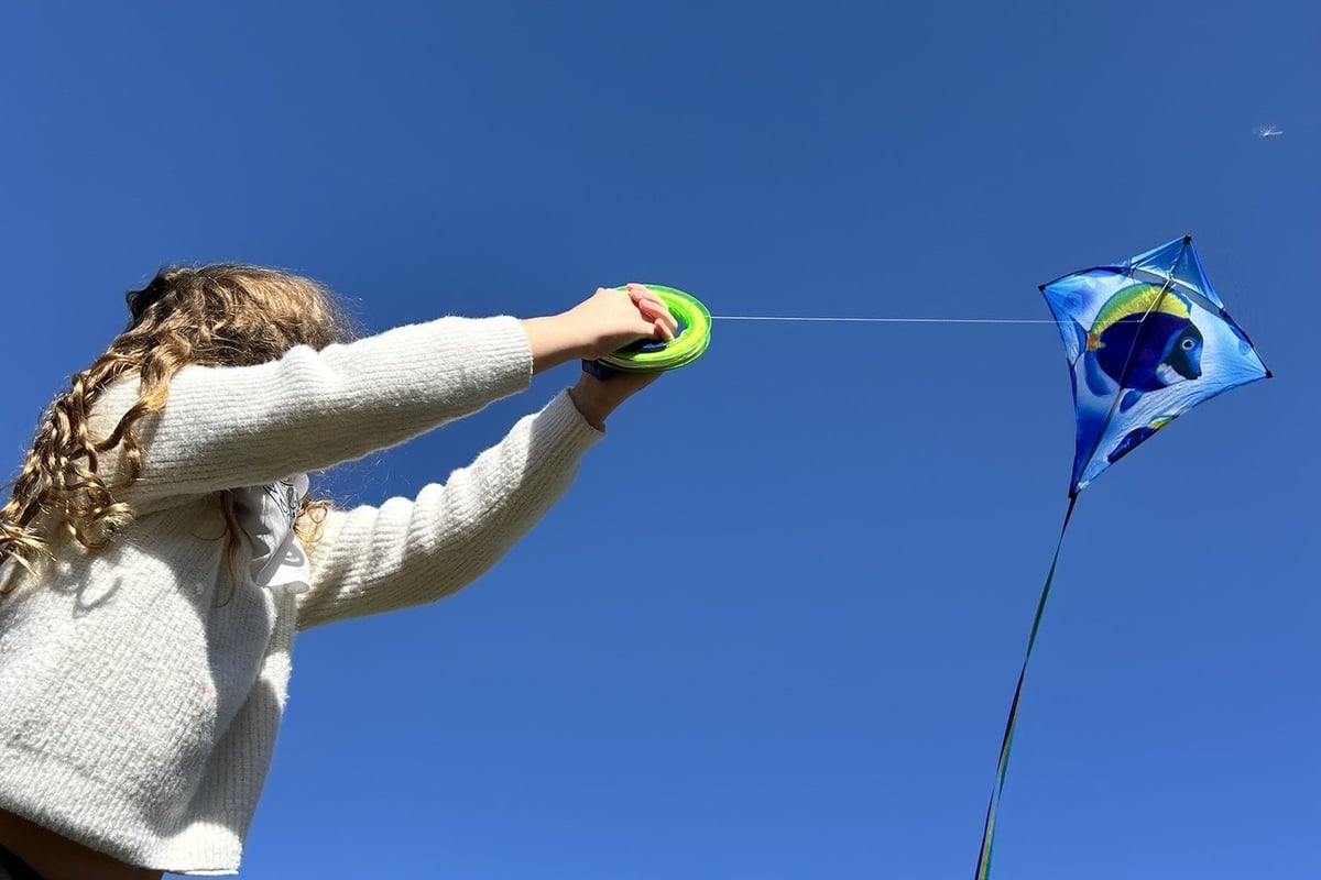Child in white sweater flying a blue diamond kite against clear blue sky.