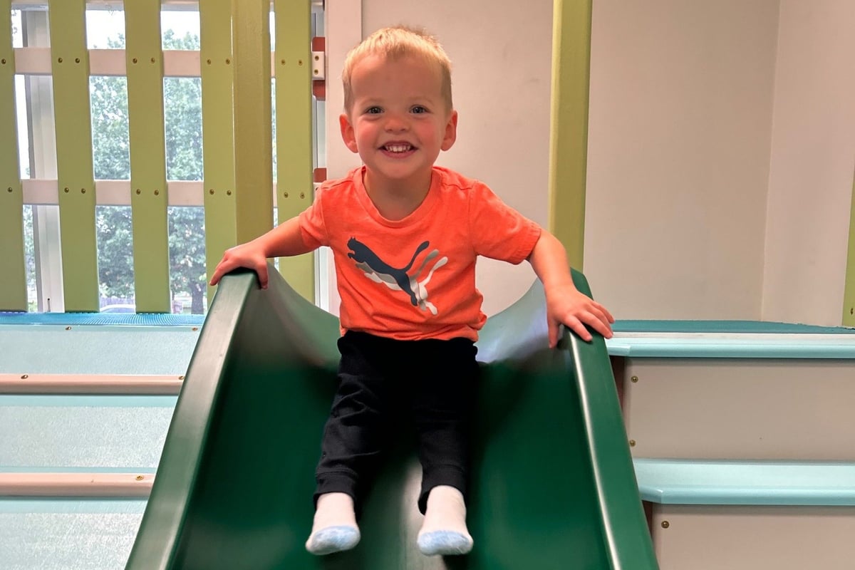 Smiling toddler in orange shirt and black pants sitting at top of green slide indoors.