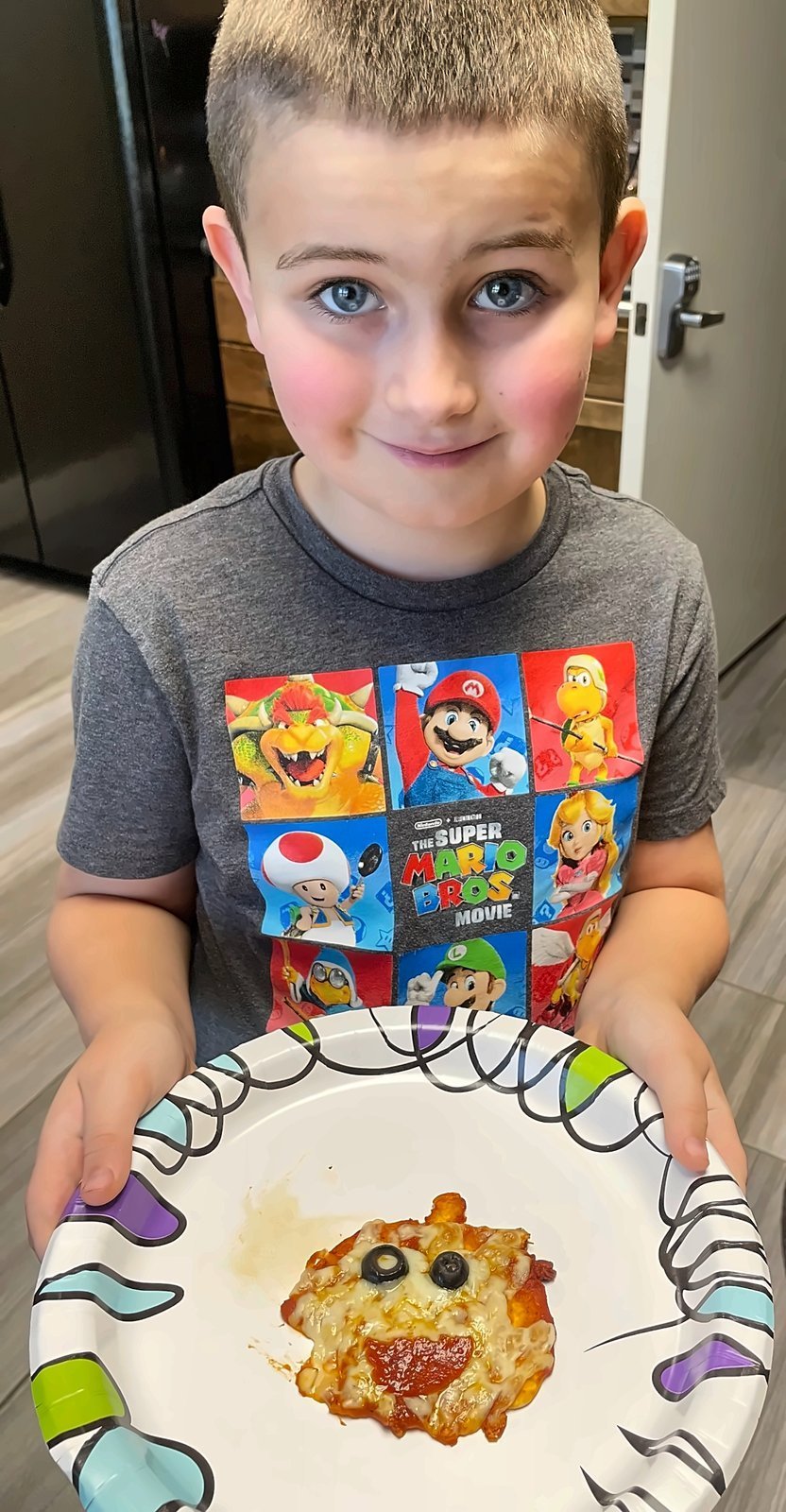 Young boy wearing a Super Mario t-shirt holding a decorated pizza face with olive eyes and pepperoni smile