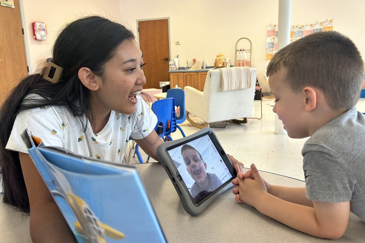 Young woman showing children a tablet device in an indoor community space, with a blue water bottle and classroom materials visible in the background.