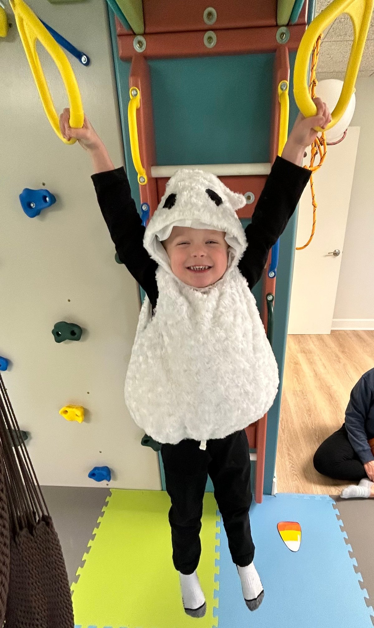 Smiling child in a sheep costume holding the yellow gym rings overhead in the sensory gym at New Heights Therapy, a pediatric occupational therapy clinic in Lawrence and Topeka, Kansas