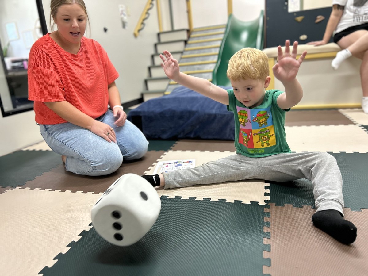 Therapist kneeling on the foam mat beside a child playing with a giant foam dice, a gross motor game in pediatric therapy